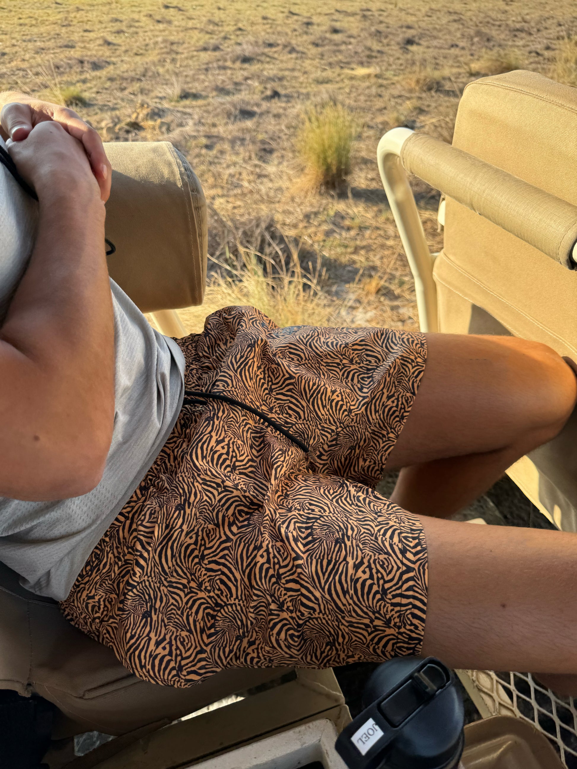 A photo features a male model sitting outdoors in tiger print swim shorts. The vibrant pattern stands out against the backdrop of his vacation in South Africa. His relaxed pose highlights the versatility of the shorts, making them suitable for both beach activities and casual outings. This stylish swimwear choice perfectly embodies the fun and adventurous spirit of summer.