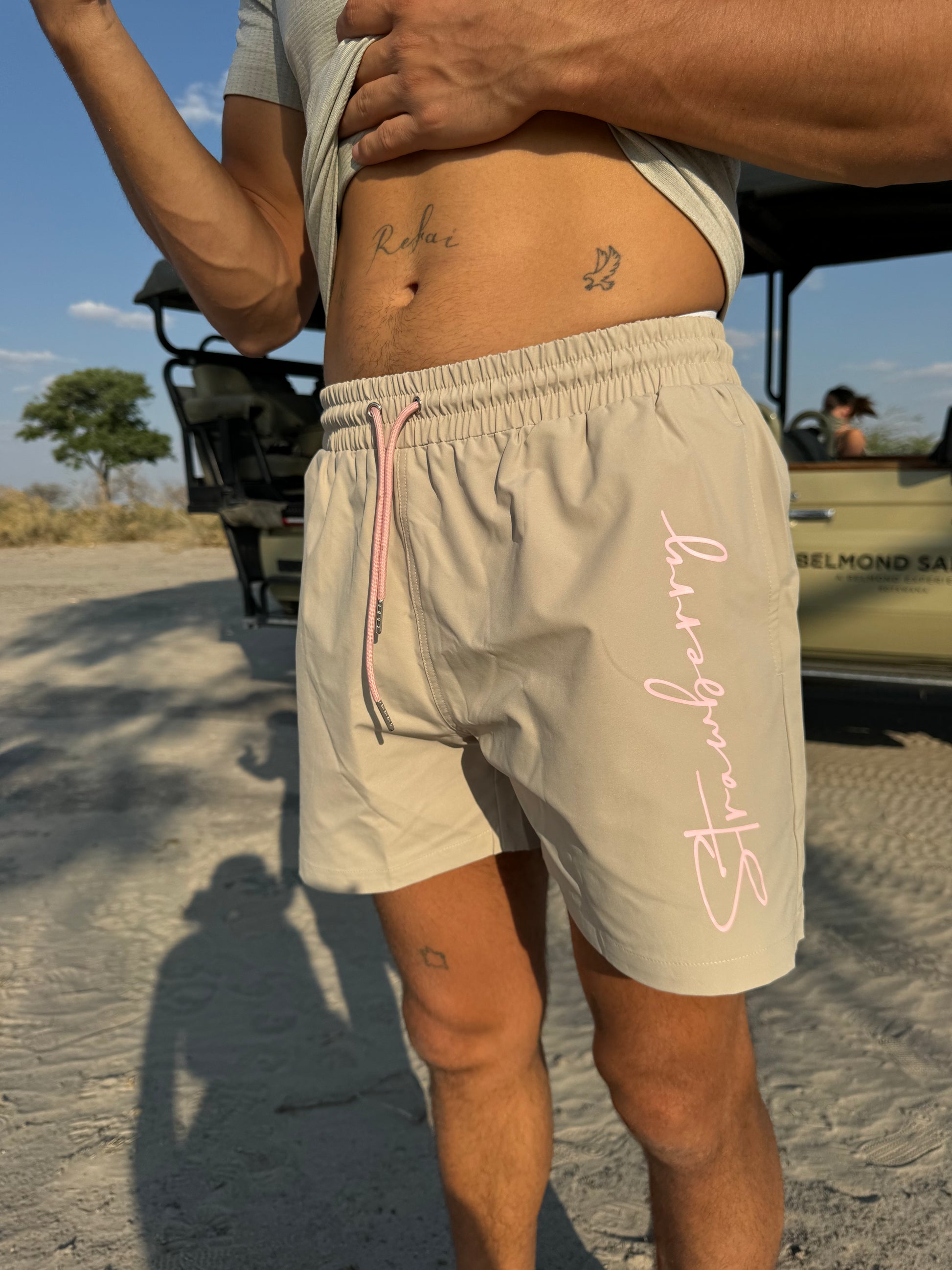 A close-up shot features a male model in versatile beige swim shorts with pink waist ties and striking "Strawberry" text detailing down the left leg. The safari backdrop is softly blurred, drawing attention to the stylish details of the outfit.