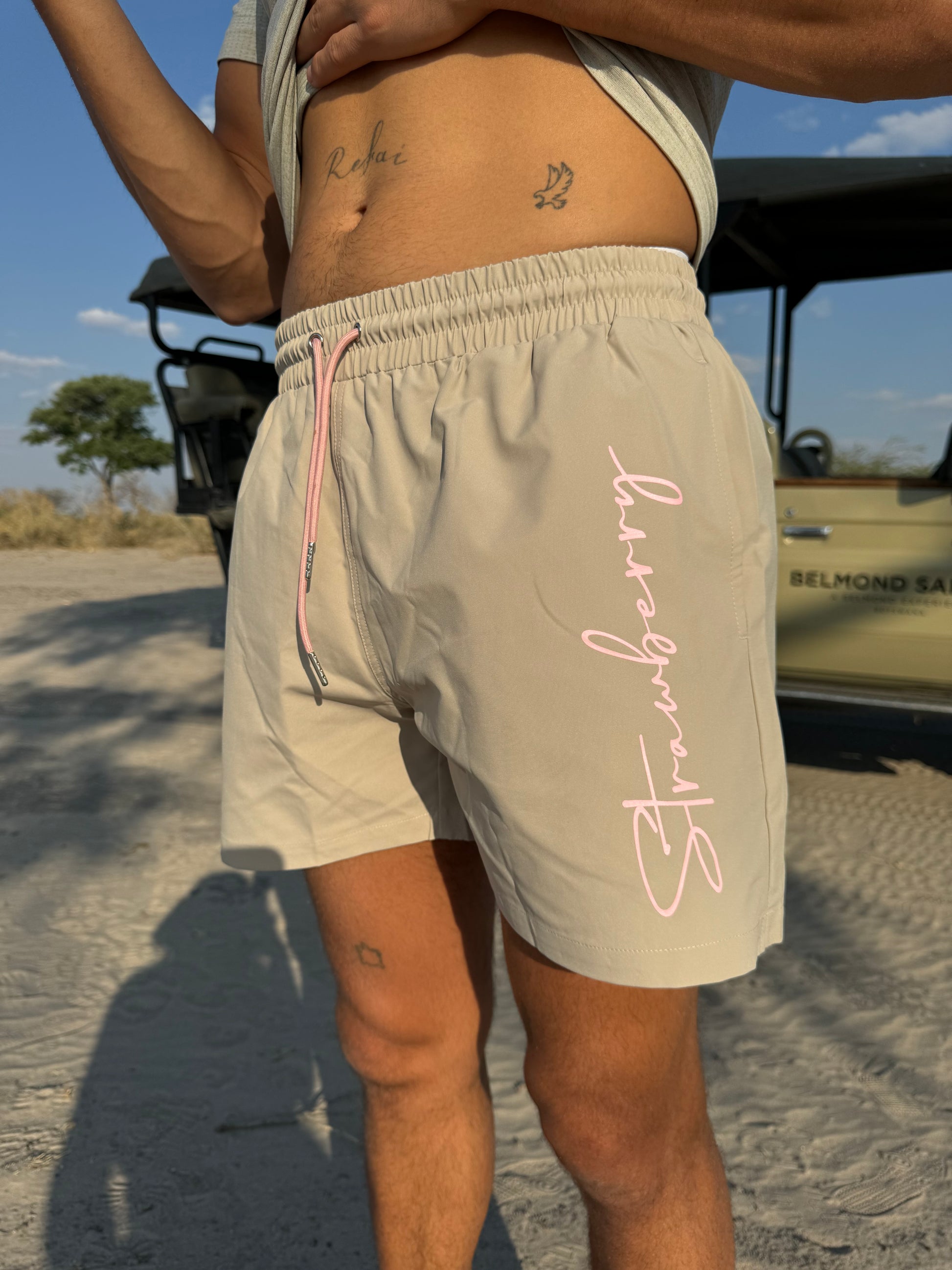 A close-up shot of a male model wearing versatile beige swim shorts with pink waist ties and bold "Strawberry" text detailing down the left leg. There is a safari backdrop softly blurred behind him, enhancing the focus on the stylish details of the outfit.