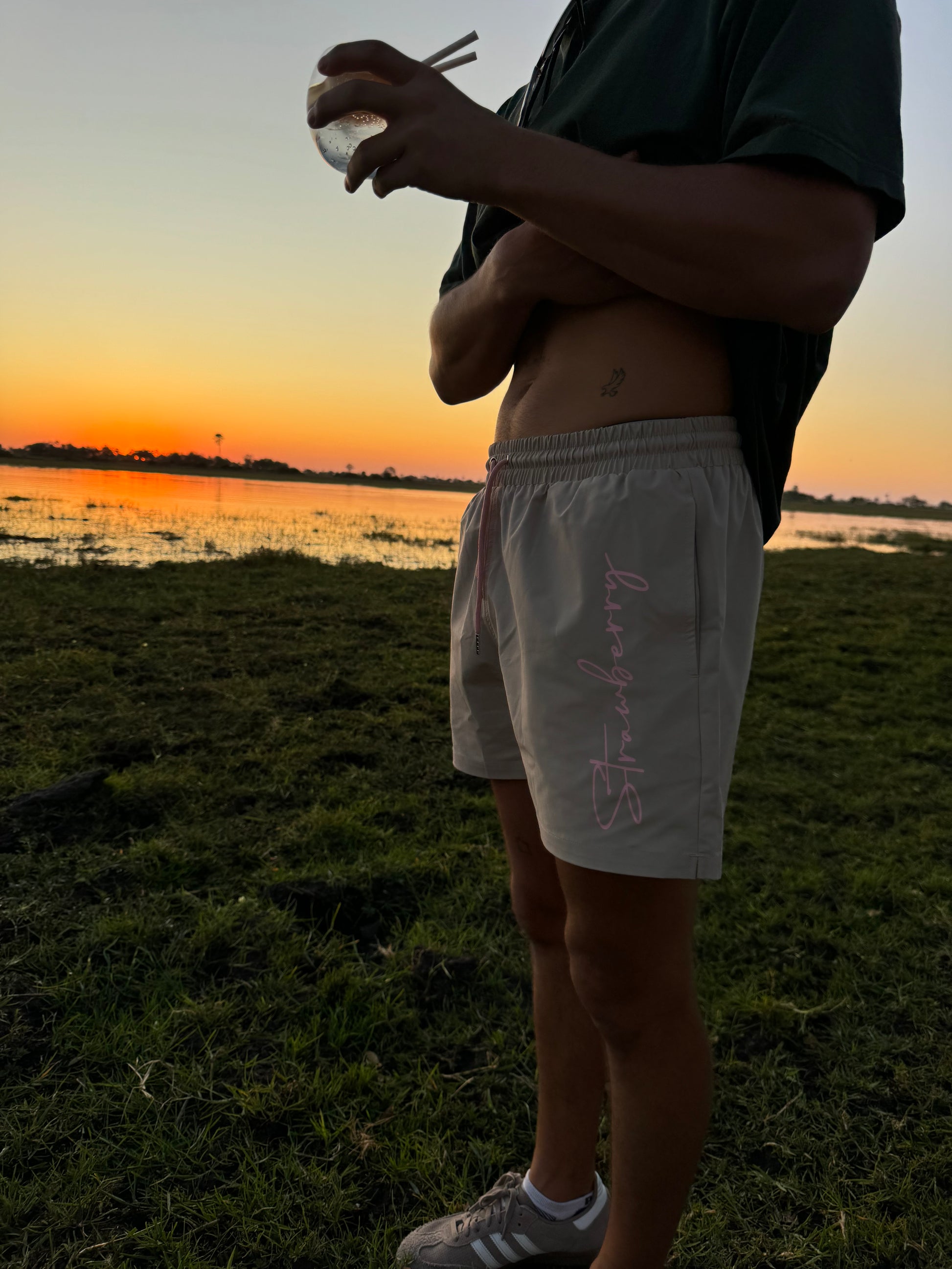 A male model stands in a wider shot, wearing versatile beige swim shorts with pink waist ties, showcasing bold "Strawberry" text detailing down the left leg. He is holding a drink, and the outdoor setting features a warm sunset in the background, enhancing the stylish look of the outfit.