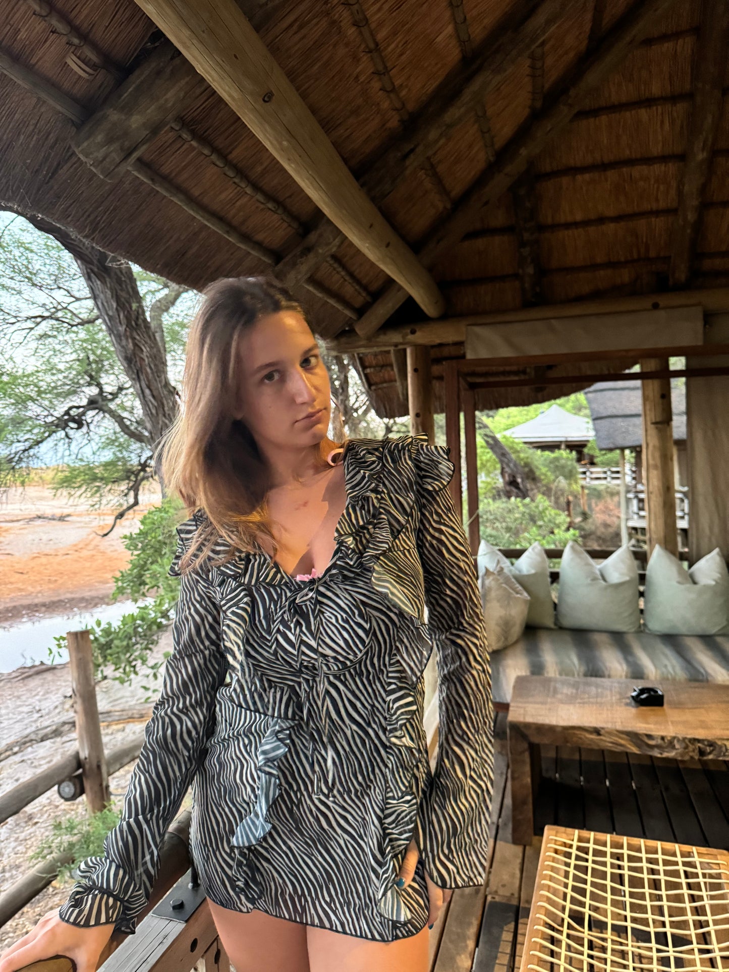 A model poses in a versatile black and white zebra stripe chiffon dress with ruffles while on vacation in South Africa. The flowing fabric drapes elegantly around her, and the vibrant patterns of the dress contrast beautifully with the natural surroundings, while warm sunlight highlights her features, creating a stylish and carefree vacation vibe.