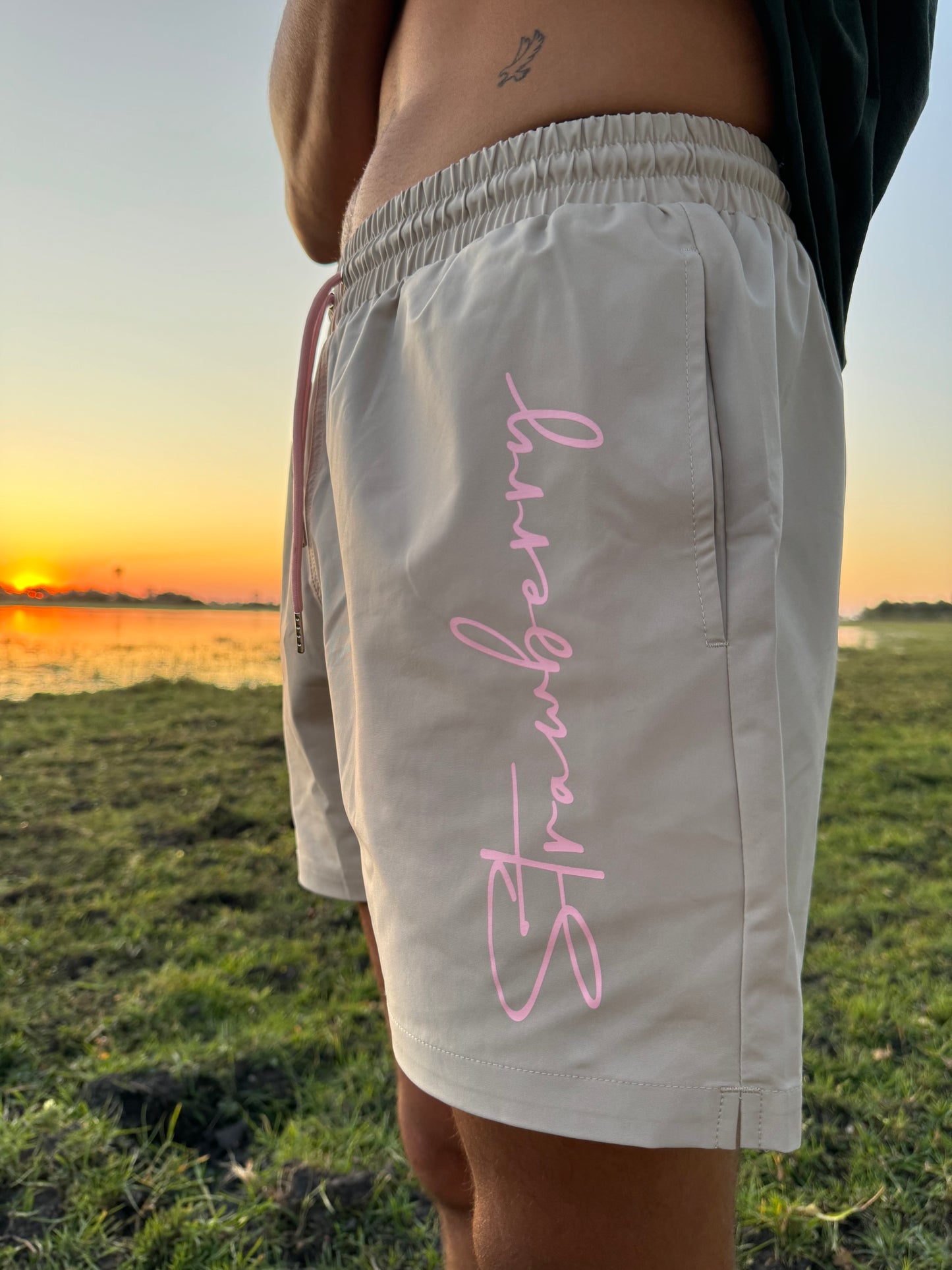 A male model poses in versatile beige swim shorts with pink waist ties, featuring "Strawberry" text detailing down the left leg. He is turned so that the focus is on the text detail and set against an outdoor backdrop with a warm sunset in the background, highlighting the stylish design.