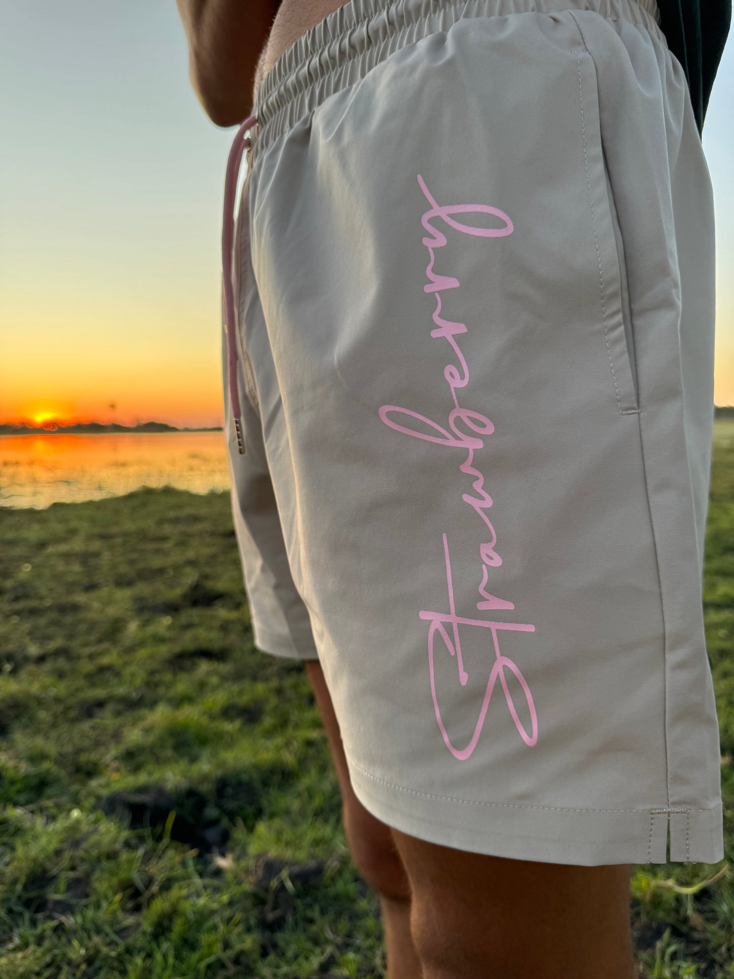 A male model showcases versatile beige swim shorts with pink waist ties, featuring bold "Strawberry" text detailing down the left leg. The focus is on the text detail, set against an outdoor backdrop with a warm sunset in the background, emphasizing the stylish design.