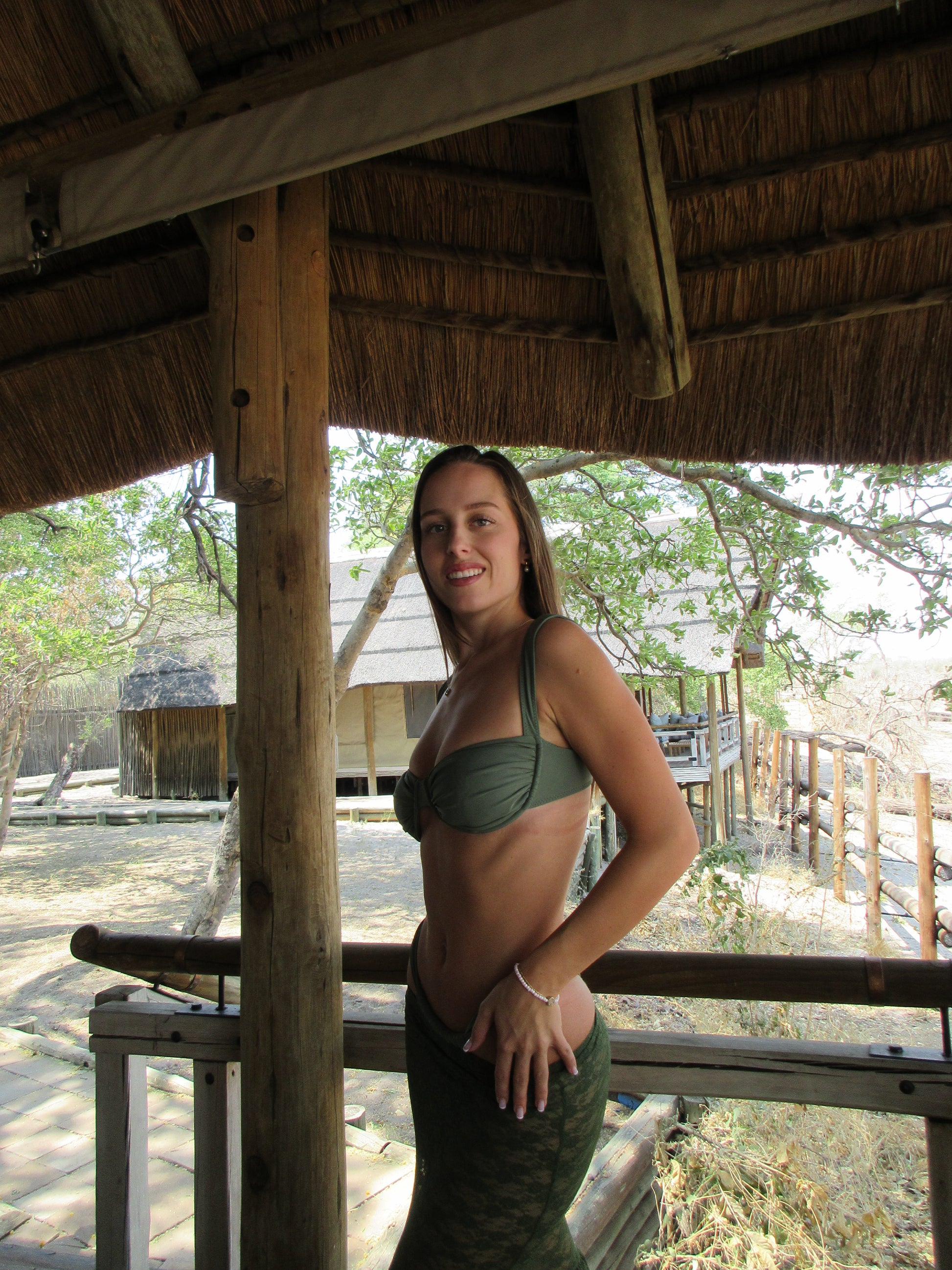 A model wearing a sage green bikini and a matching layered lace skirt poses under some shade on her vacation in South Africa. The soft light filters through, creating a gentle ambiance that highlights the textures of her outfit. She stands confidently, while the scenery around her adds to the tranquil atmosphere, making for a perfect summer scene.