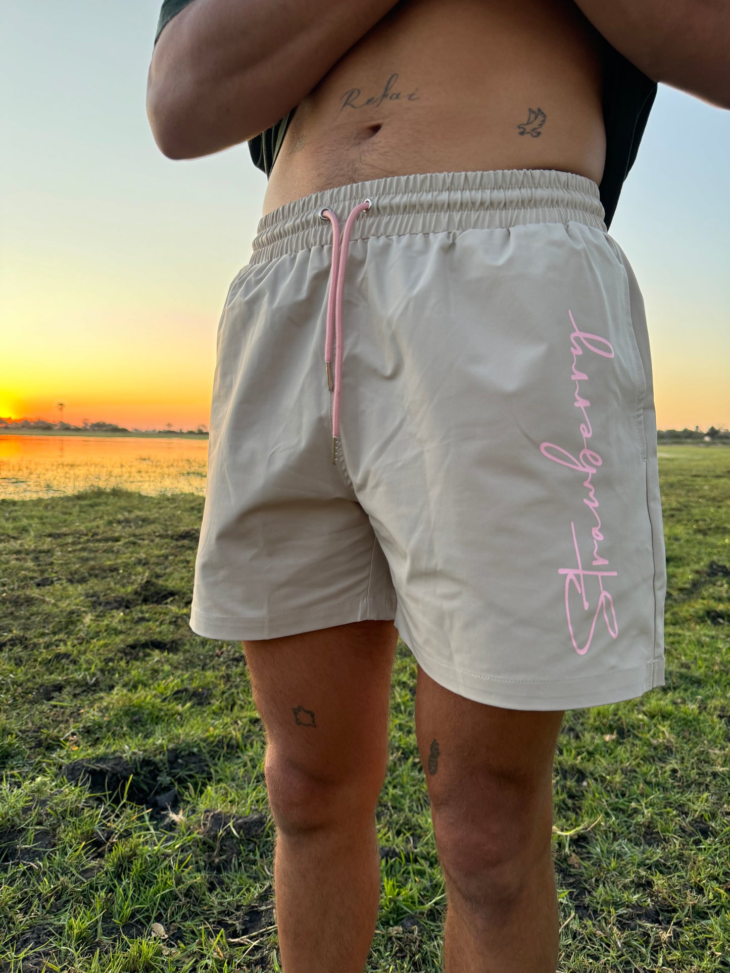 A male model poses in versatile beige swim shorts with pink waist ties and "Strawberry" text detailing down the left leg. Set against an outdoor backdrop, the scene is illuminated by a warm sunset, enhancing the stylish look of the shorts.